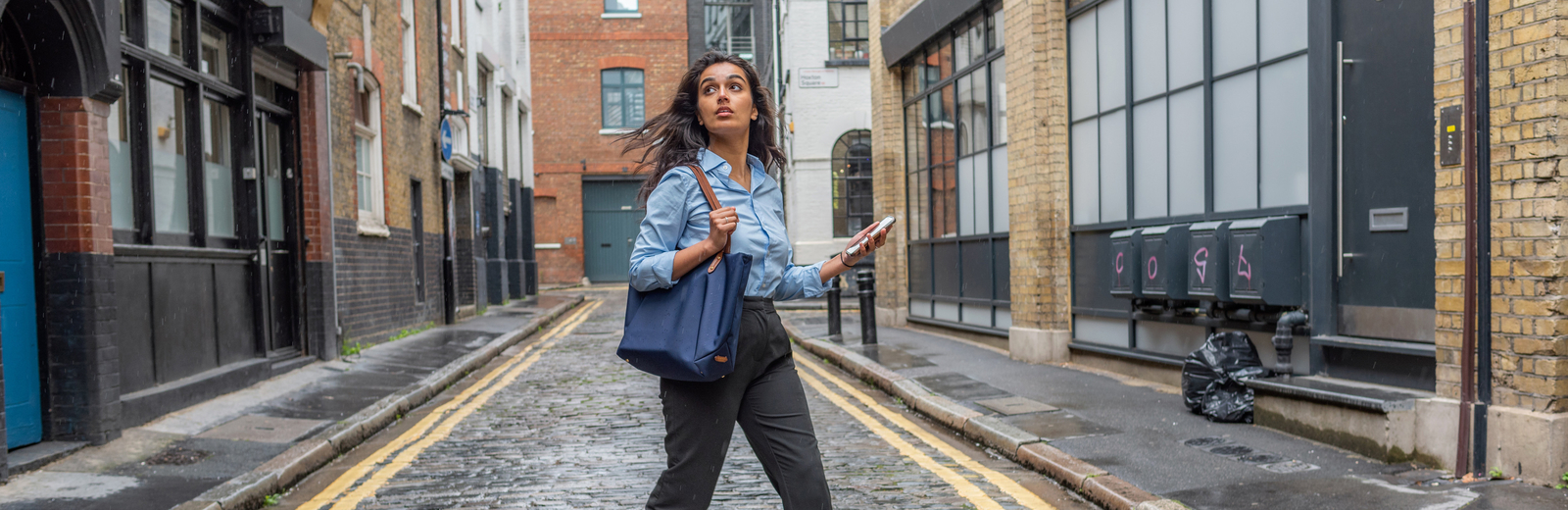 Female student looking around as she walks through a street in Shoreditch, smartphone in hand.