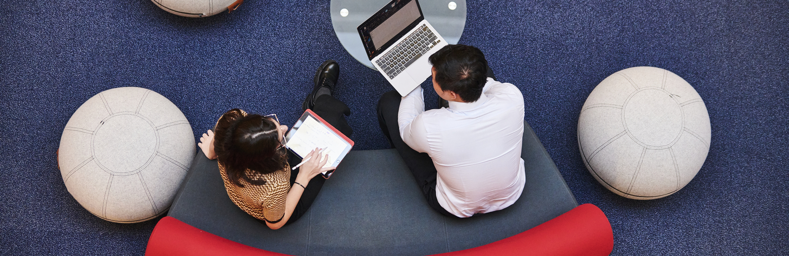 Looking down from above - a woman and man sitting together on a curved atrium sofa. The man looks at a laptop, while a woman holds a tablet and stylus.