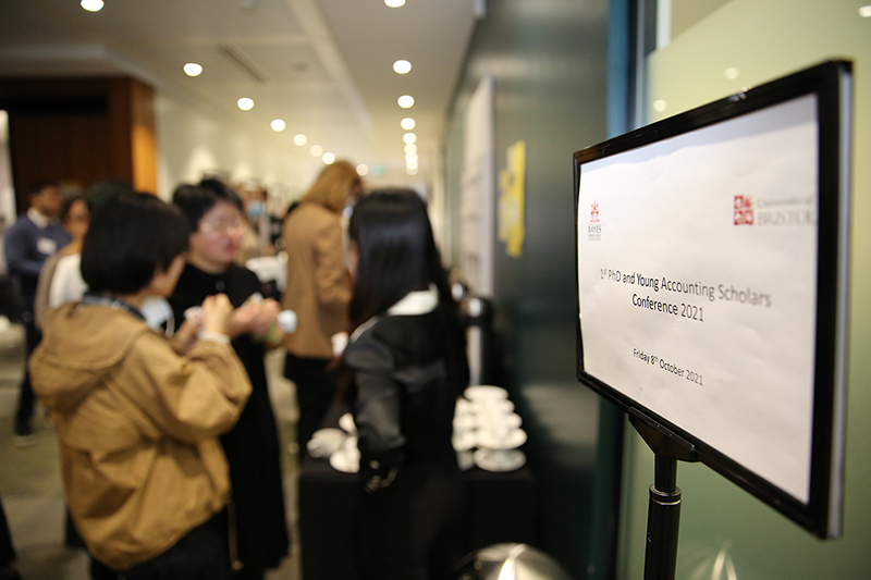Event signage "First PhD and Young Accounting Scholars Conference 2021", with out-of-focus group of attendees having a conversation in the background.