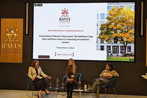 Image of speaker Natalie Bonnick, Dr Stephanie A Baker and Daniella Isaacs sitting on chairs on stage at Bayes Business School in an amphitheatre lecture room. Behind them is a large screen with the event title