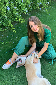 Melisa Lara Denizoglu sits on the grass, smiling and playing with a small dog. She is dressed in a green outfit with orange-accented sneakers. 