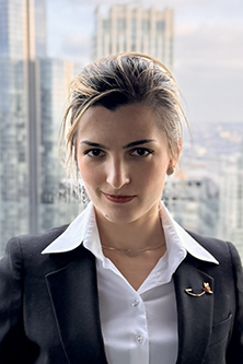 A professional headshot of Diana Calmic with blonde hair, wearing a black blazer and white collared shirt, standing in front of a cityscape with tall buildings. She has a confident expression and a subtle smile.