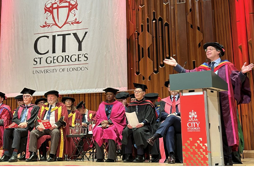 People in formal academic gowns sit on a stage listening to a man talking.