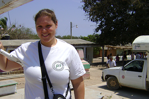 Young woman standing outside before some sheds and a van, wearing a white tshirt with a PBI logo, and carrying camera cases.