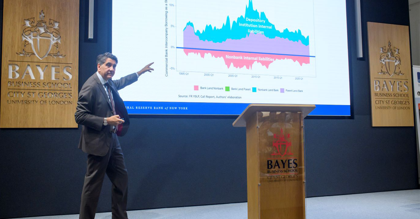A middle-aged white man speaking on a stage and pointing to a screen with data on it