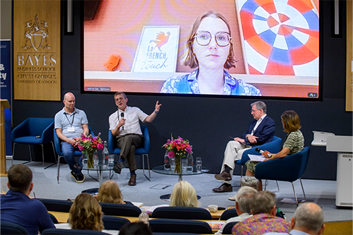 Four people sit on a stage while a woman is on a screen behind them