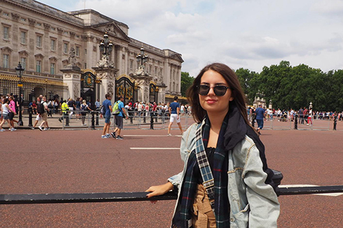 Melisa Lara Denizoglu stands in front of Buckingham Palace in London. She is wearing a denim jacket and a plaid scarf, with sunglasses on. The background shows the palace gates and a crowd of tourists.