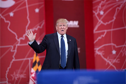 Donald Trump waves at a Conservative Political Conference in the USA. He wears a dark suit and blue tie, and is against a red backdrop with white outlines in the shape of the US states. Ahead of him is a blue lectern