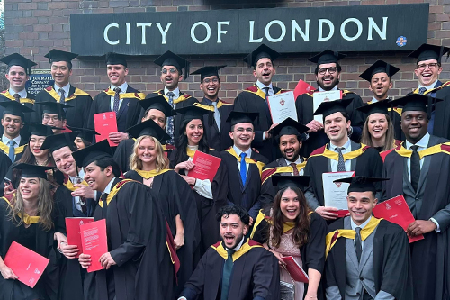 A large group of diverse graduates in black and gold academic gowns and mortarboards celebrate their graduation outside a building with a "City of London" sign. Many are holding red diplomas and smiling joyfully.