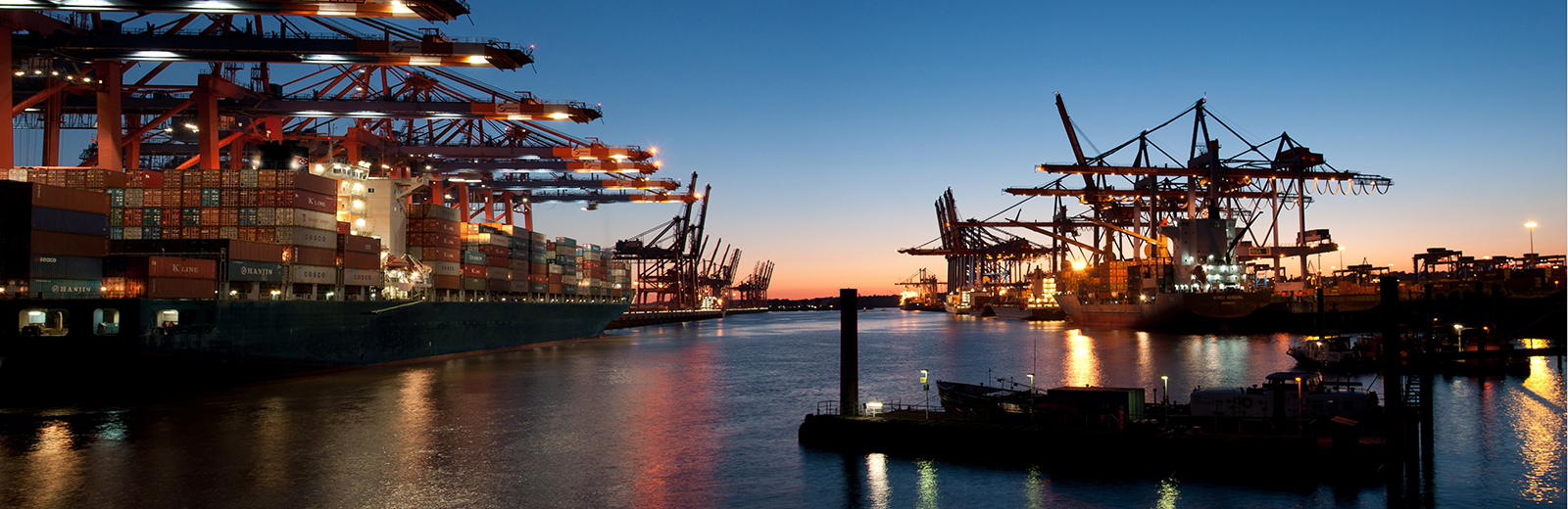 Hamburg harbour - ships loaded with shipping containers at sundown, with lights reflected on the sea.