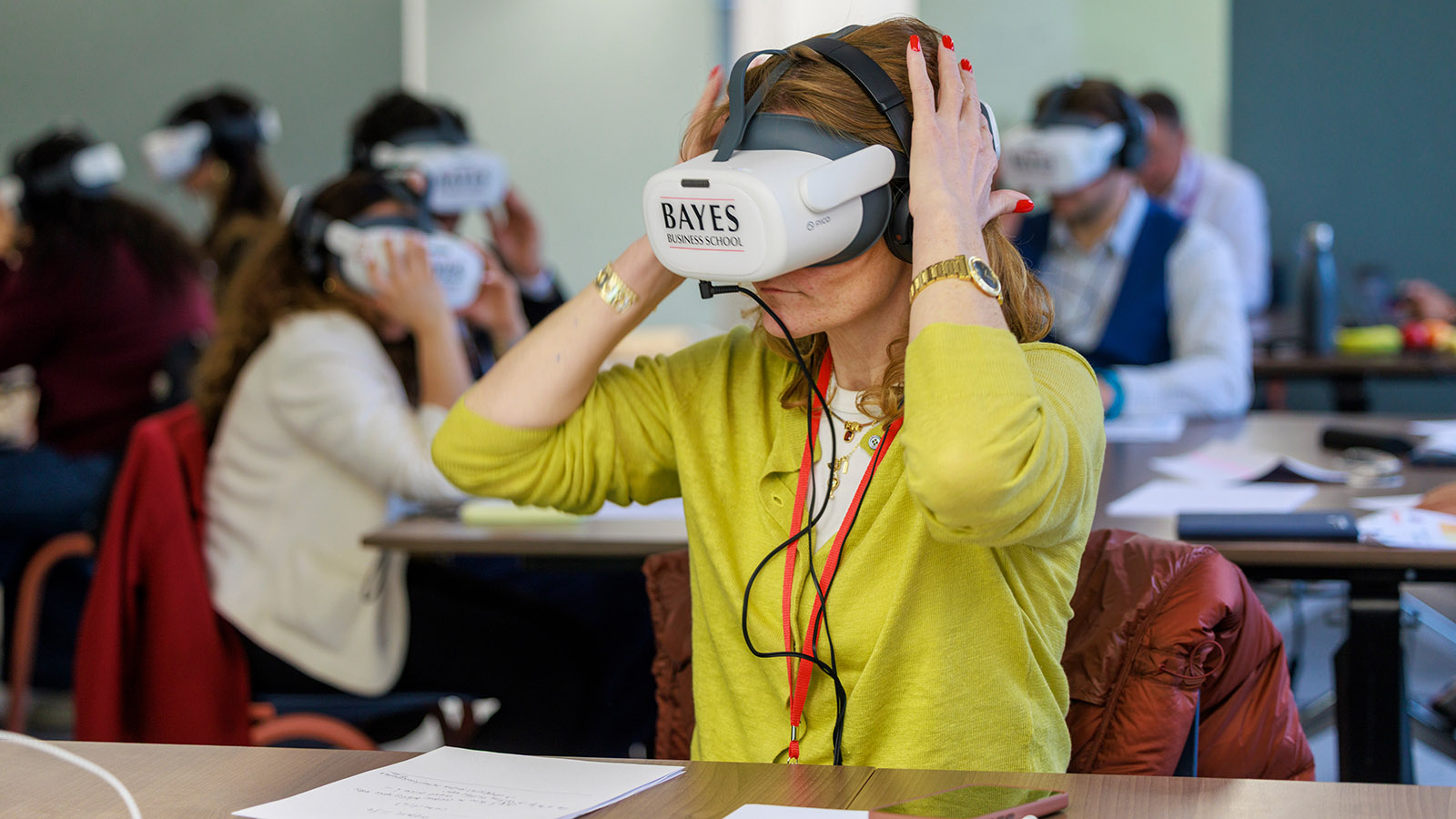 A woman supports a VR headset bearing the Bayes logo on her head. Behind her other people in a classroom setting are also wearing headsets.