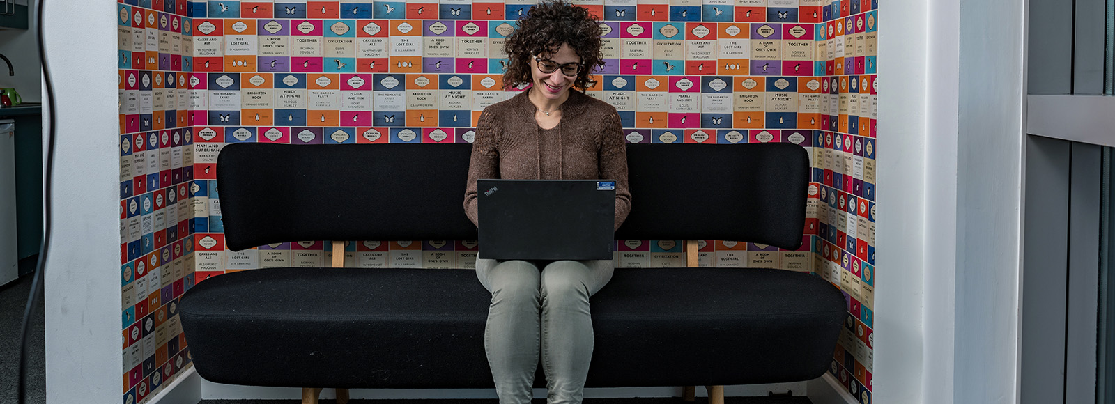 Smiling woman in casual clothes, sat on a sofa in the university, typing something on her laptop.