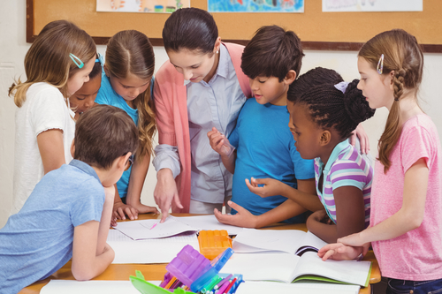 A teacher stands among a diverse group of schoolchildren at a desk and points to an open notebook. Behind them, you can see a blurry background with children's drawings