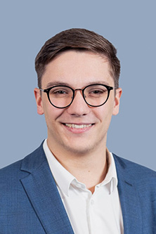 Eric Scheerbarth wearing round black glasses, a white dress shirt, and a blue suit jacket, smiling in front of a solid light blue background.
