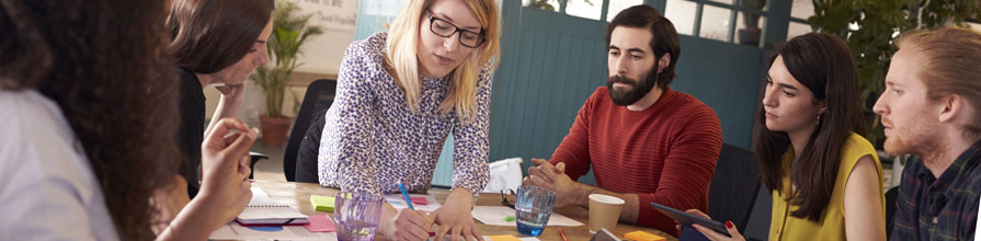A female speaker leads a small group of professionals through a workshop around a table.