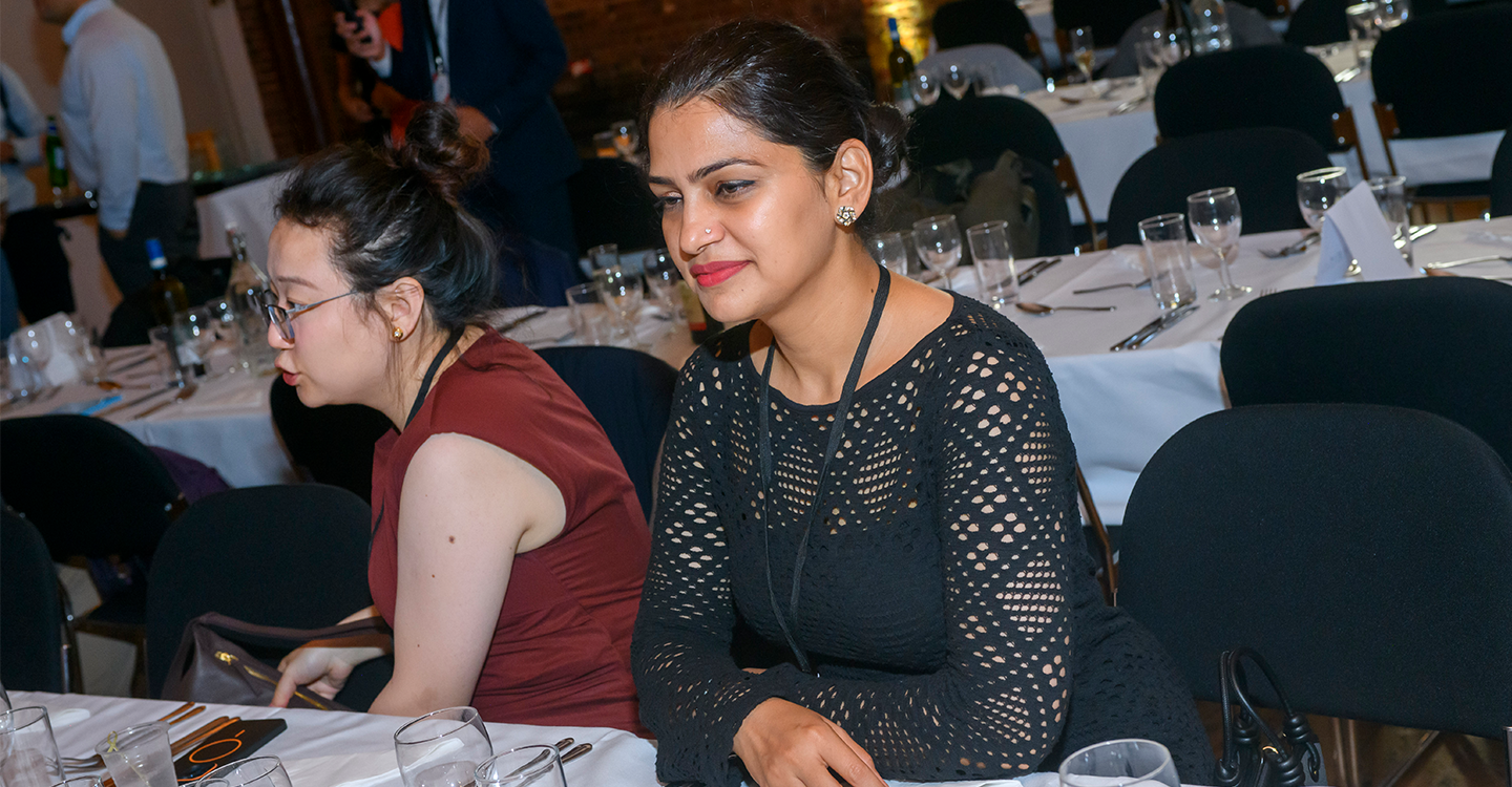 Two women are speaking while sitting at a table in a crowded room