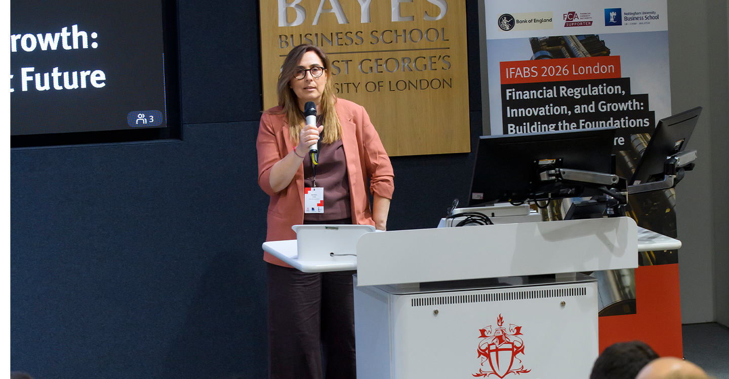 A white woman speaks with a microphone in front of a Bayes crest of arms