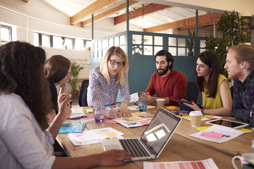 A female speaker leads a small group of professionals through a workshop around a table.