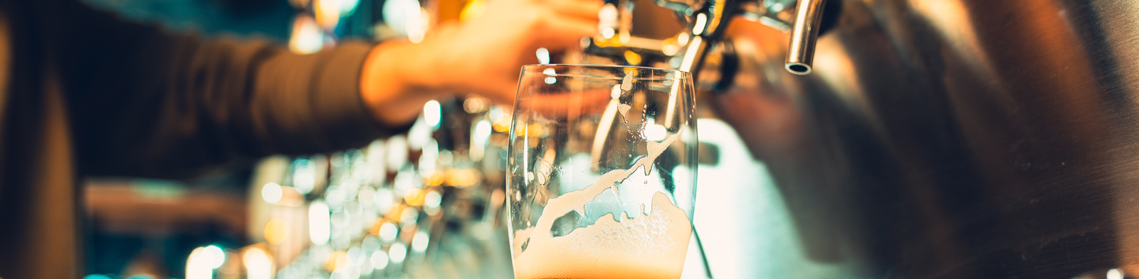 A young bartender pours beer from a tap into a glass in a busy bar.
