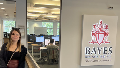 Melisa Lara Denizoglu is standing next to a sign that reads "Bayes Business School, City University of London." Behind her, there is a glass wall revealing a classroom with people working on computers. The room is well-lit with natural light coming through the windows.