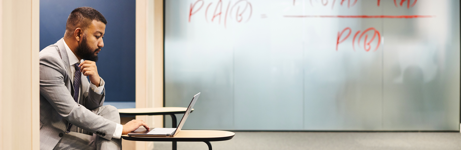MBA student sitting with laptop in Finsbury Square building, Bayes Theorem printed large on the internal frosted window behind him.