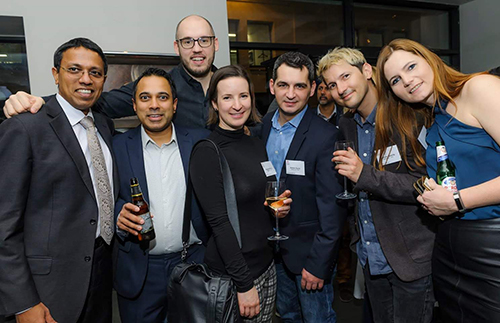 Group photo of men and women with wine glassed and beer bottles in their hands at an evening social event.