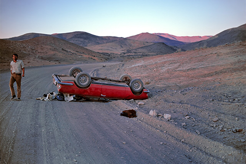 A man on a deserted road looks out at the camera standing near his overturned red car