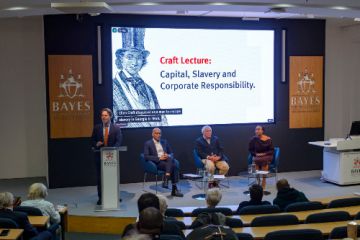 Speakers on stage in a Bayes lecture theatre, under a screen displaying the lecture title slide.