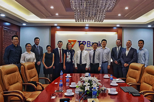 Group photo of professional women and men in a luxurious looking board room with flower arrangements on the table, standing before a large sign on the wall saying 'TP Bank'.