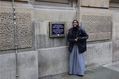 A young woman stands next to a blue plaque marking the place where Mary Harris Smith, the world's first female chartered accountant, had her office.