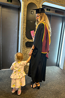 Caroline Hamren at graduation in the Barbican. She's wearing graduation robes and standing before the lifts with her young daughter.