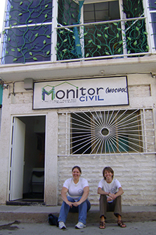 Two women sitting on the side of the road outside a building with a sign saying Monitor Civil (Mocipol). The upper floor windows are covered with decorative iron-work vines